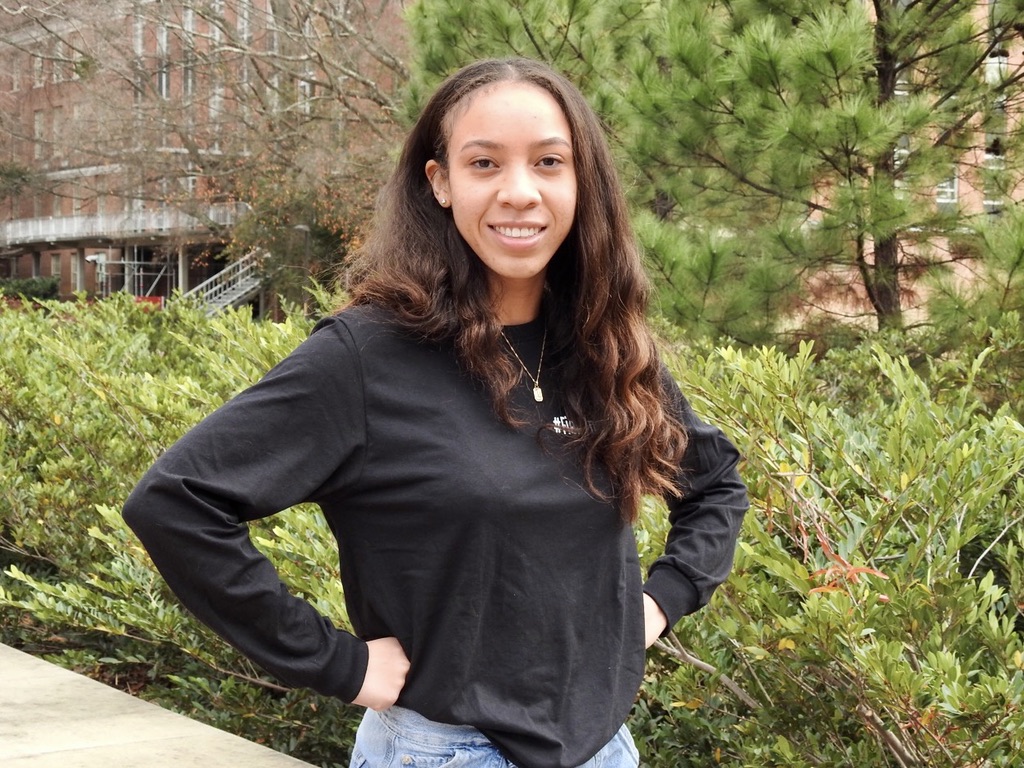 Kayla Stukes, M.S: Volunteer. Kayla has dark long, wavy hair and is wearing a black long-sleeve shirt and light blue jeans. Their hands are placed on their hips. In the background, there is greenery, including a tree with dense foliage and some bushes. The setting is near a building, part of the structure is visible on the left side of the image.
