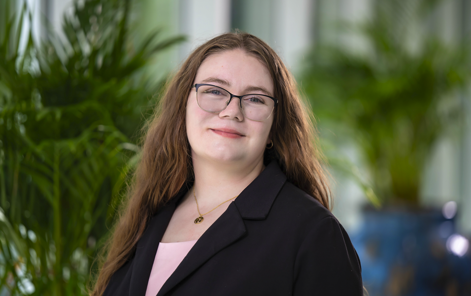 Grace Albertson is the Social Media and Communications intern. She has long, wavy brown hair and is wearing black-rimmed glasses. She is wearing a black blazer over a light pink top. Around the neck, there is a thin gold necklace with a small, oval-shaped pendant. The background features a blurred setting with green foliage.